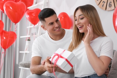 Photo of Man presenting gift to his girlfriend in room decorated with heart shaped balloons. Valentine's day celebration