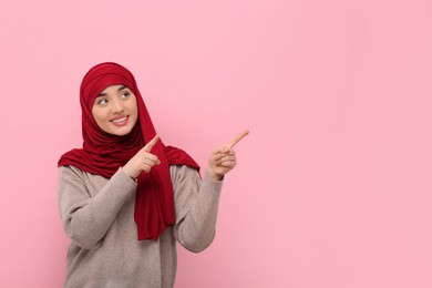 Muslim woman in hijab pointing at something on pink background, space for text