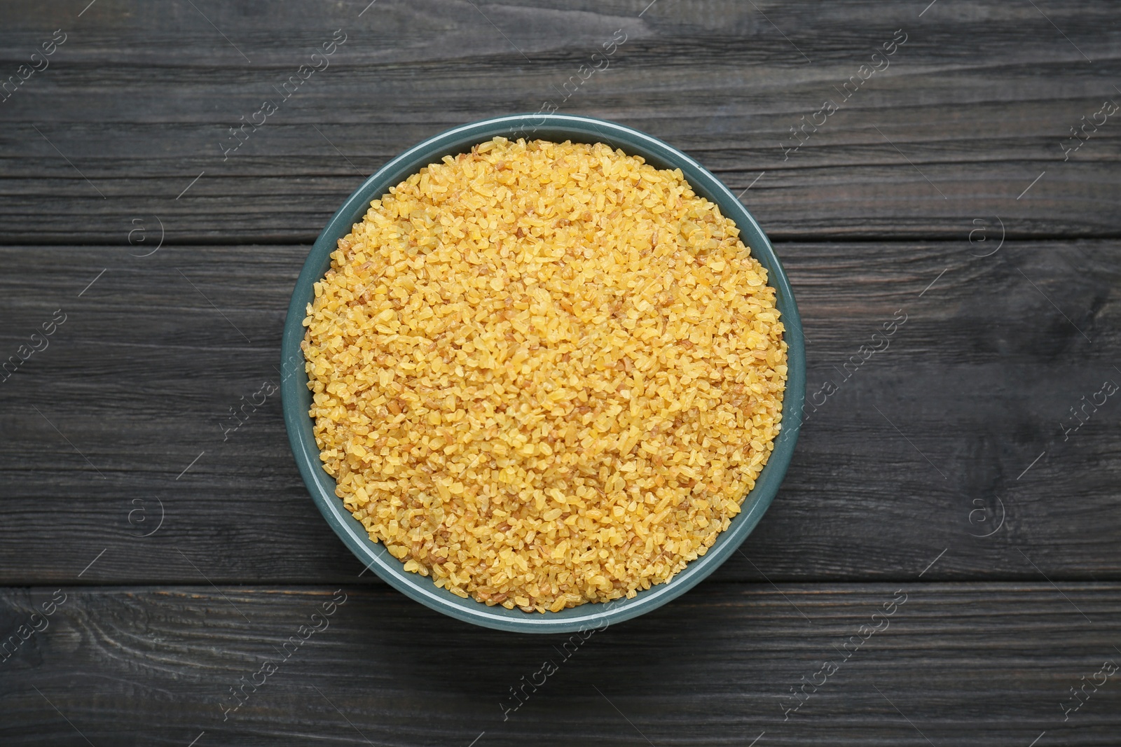 Photo of Bowl of uncooked bulgur on black wooden table, top view