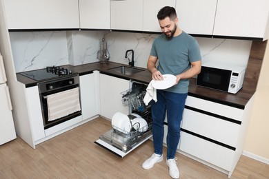 Smiling man wiping plate near open dishwasher in kitchen