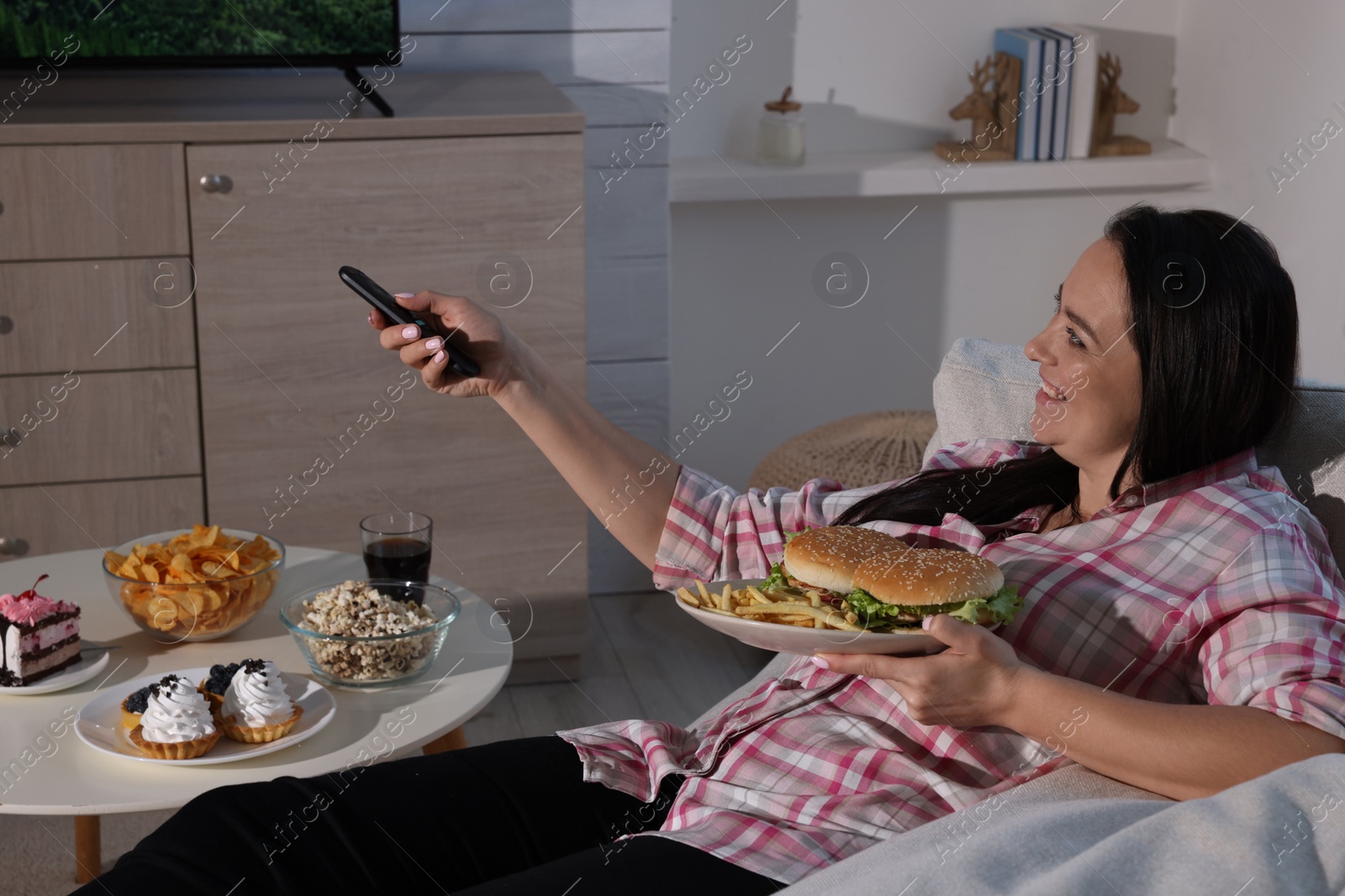 Photo of Happy overweight woman with unhealthy food watching TV on sofa at home