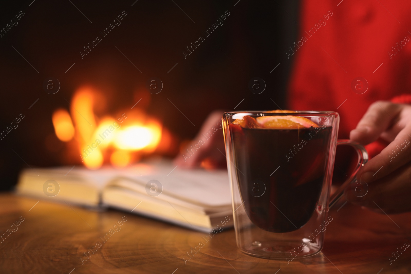 Photo of Woman with mulled wine reading book near fireplace indoors, closeup. Winter vacation