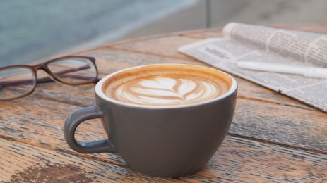Cup of delicious coffee, eyeglasses and newspaper on wooden table, closeup