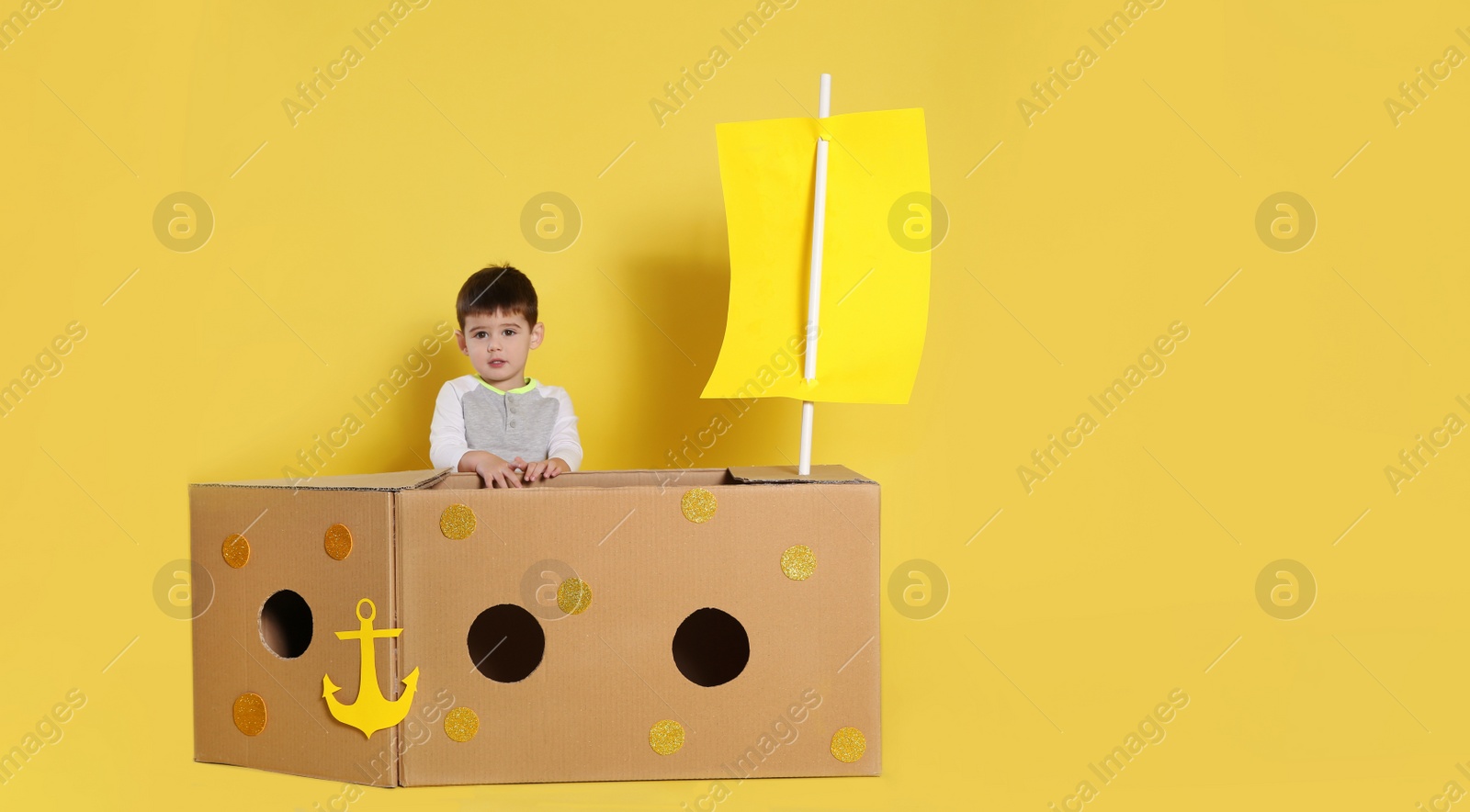 Photo of Cute little child playing with cardboard ship on yellow background