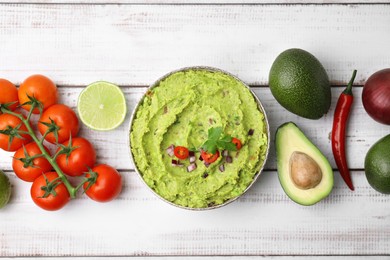 Bowl of delicious guacamole and ingredients on white wooden table, flat lay