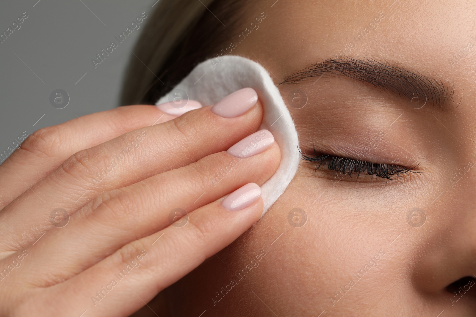 Photo of Beautiful woman removing makeup with cotton pad on grey background, closeup