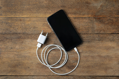 Smartphone and charging cable with adapter on wooden table, flat lay