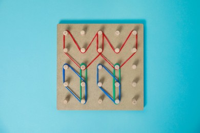 Photo of Wooden geoboard with flower shapes made of rubber bands on light blue background, top view. Educational toy for motor skills development
