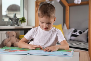 Photo of Cute little boy reading book at table in room