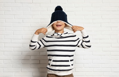 Photo of Cute little boy in hat and sweater near white brick wall. Winter season
