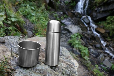 Photo of Cup and vacuum flask on rock near mountain waterfall. Space for text