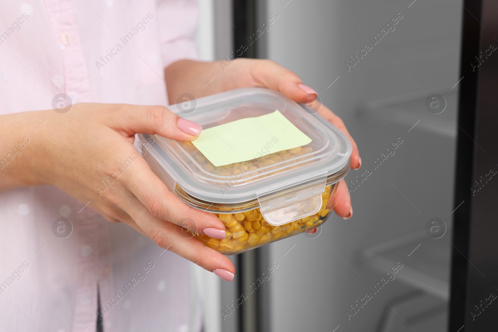 Photo of Woman holding container with tasty corn kernels near fridge, closeup. Food storage