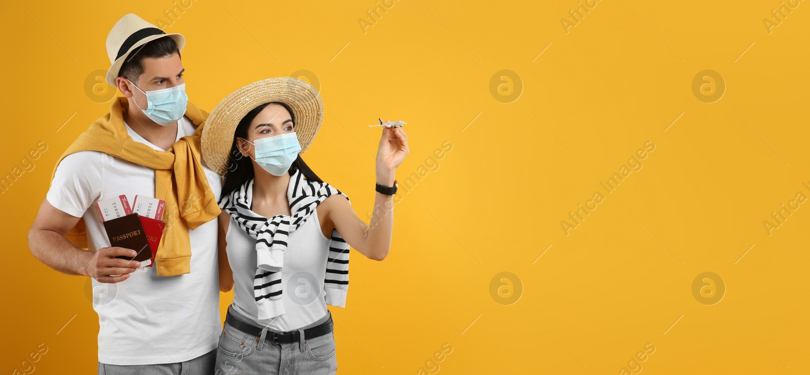 Photo of Couple of tourists in medical masks with tickets, passports and toy plane on yellow background. Travelling during coronavirus pandemic