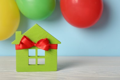 Photo of Wooden house model with red bow and balloons on light blue background, space for text. Housewarming party