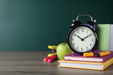 Photo of Alarm clock and different stationery on wooden table near green chalkboard, space for text. School time