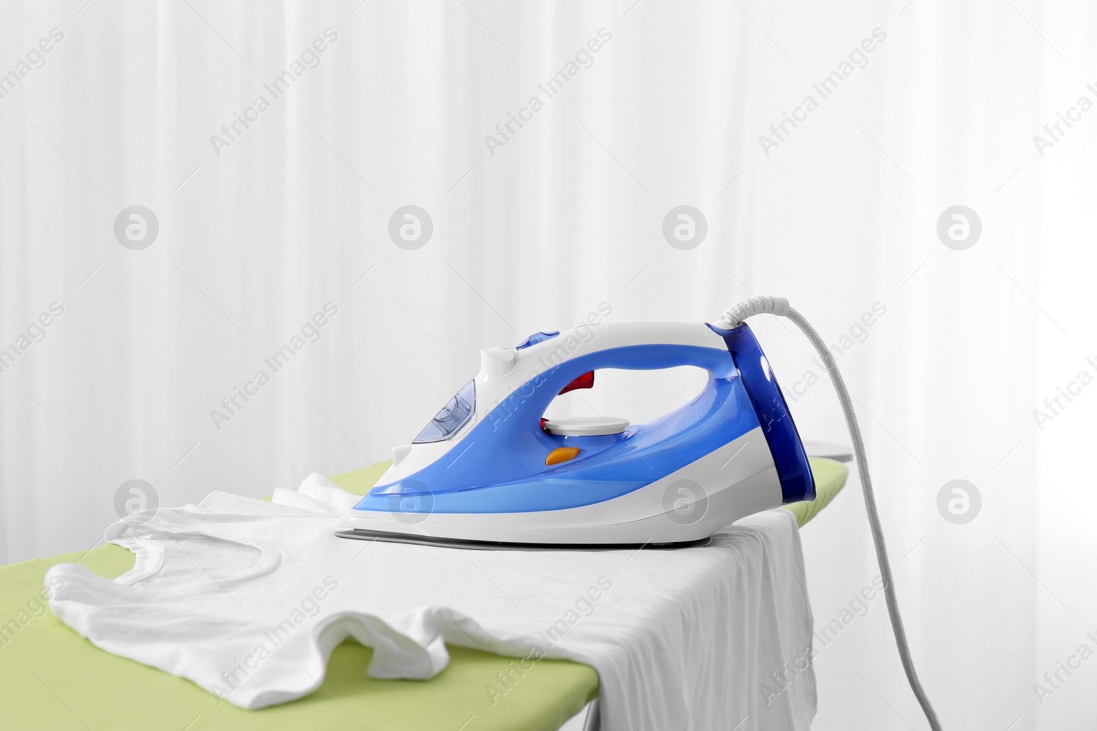 Photo of Modern electric iron and t-shirt on board indoors. Laundry day