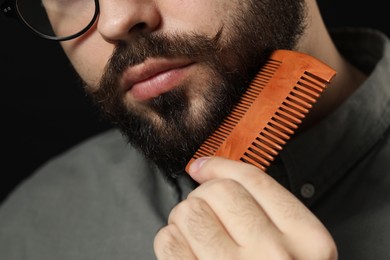 Photo of Handsome young man combing beard on black background, closeup