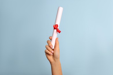 Student holding rolled diploma with red ribbon on light blue background, closeup