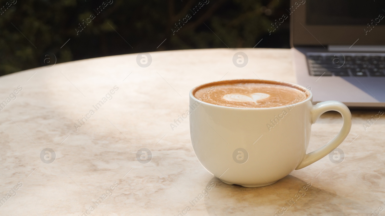 Photo of Cup of delicious coffee and laptop on beige marble table outdoors, space for text