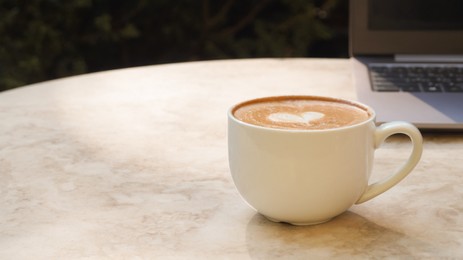 Photo of Cup of delicious coffee and laptop on beige marble table outdoors, space for text