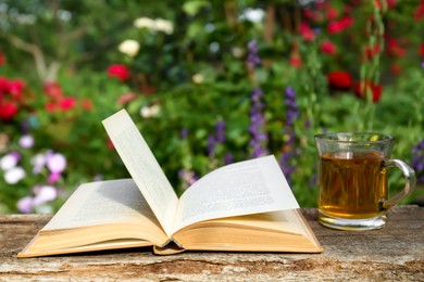 Photo of Open book with glass cup of tea on wooden table in garden