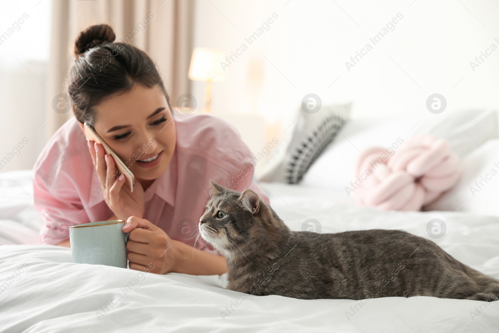 Photo of Young woman with cup of coffee talking on phone while lying near cute cat in bedroom. Pet and owner