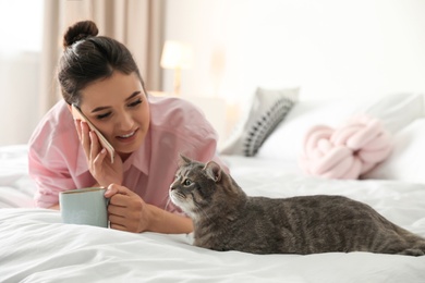 Young woman with cup of coffee talking on phone while lying near cute cat in bedroom. Pet and owner