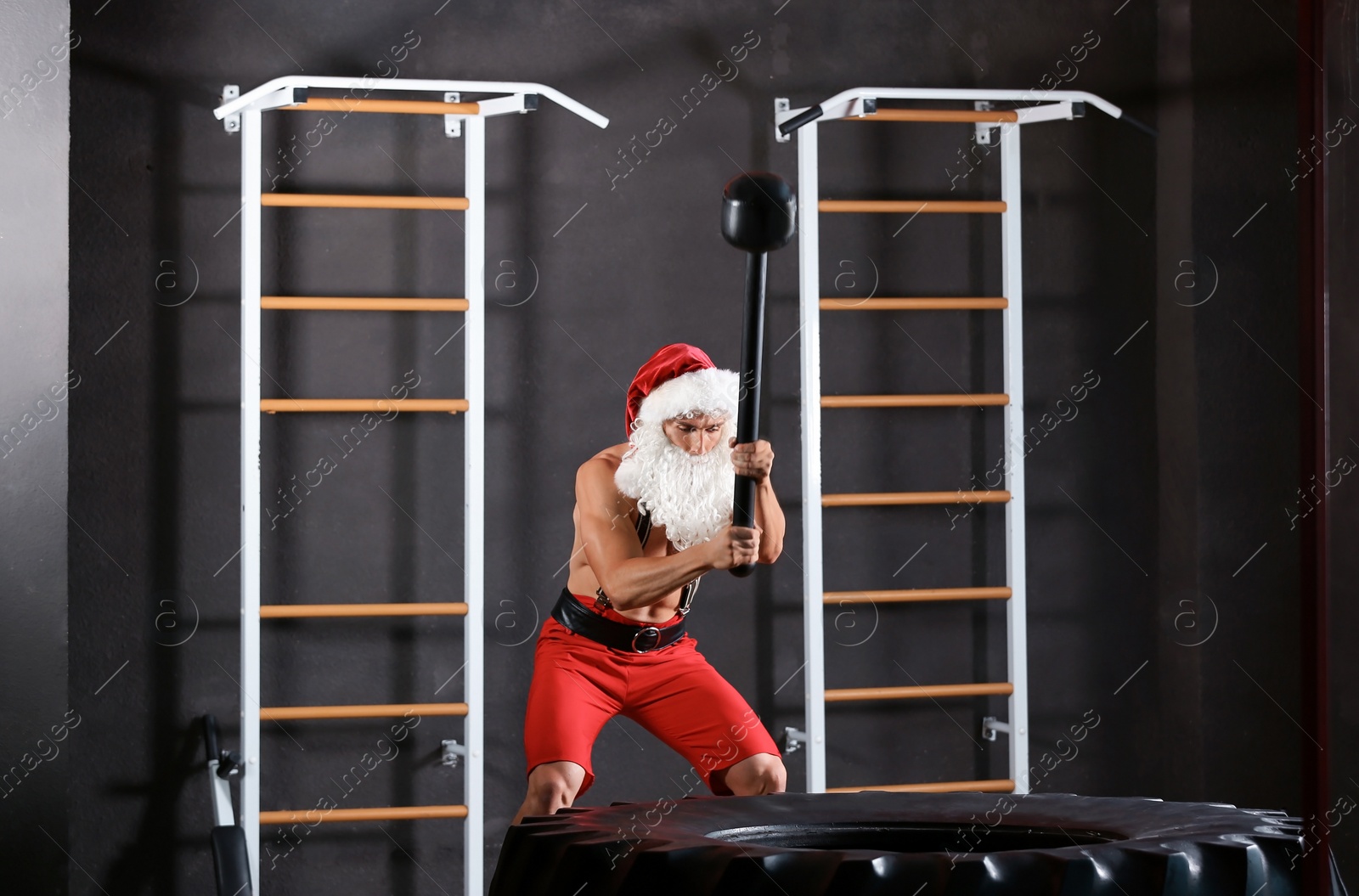 Photo of Young muscular man in Santa costume hitting heavy tire with hammer at gym