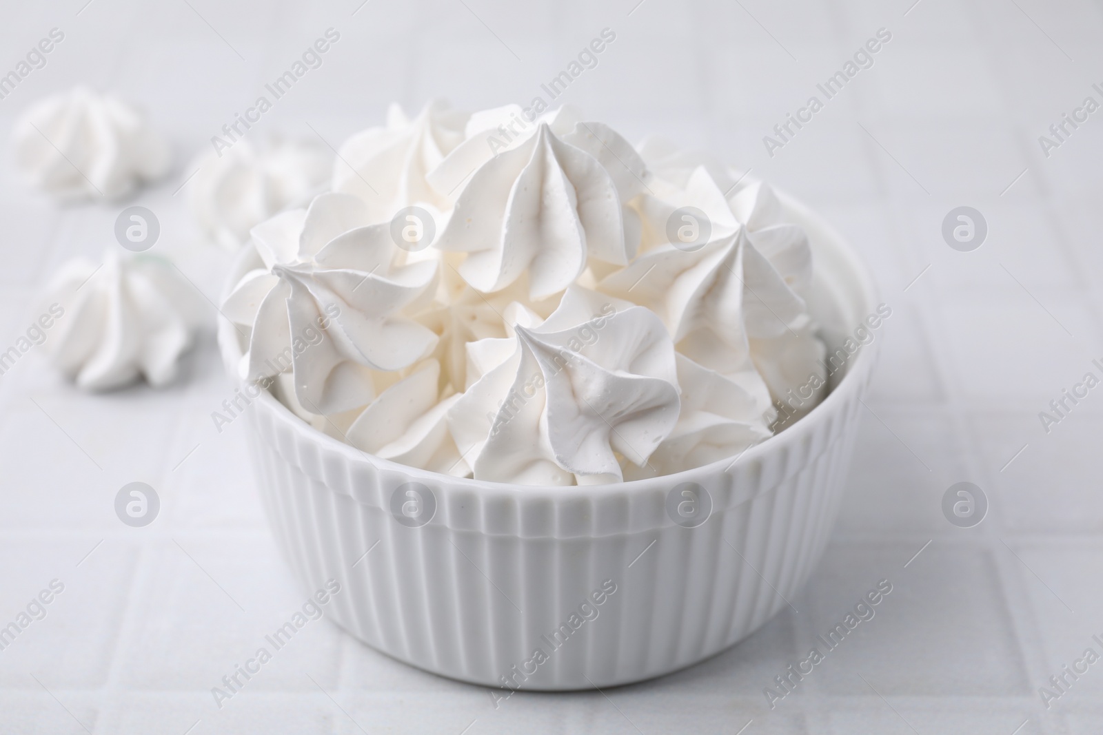 Photo of Delicious meringue cookies in bowl on white tiled table, closeup