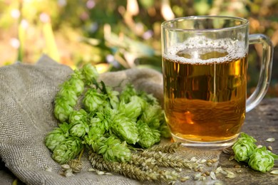 Photo of Mug with beer, fresh hops and ears of wheat on wooden table outdoors