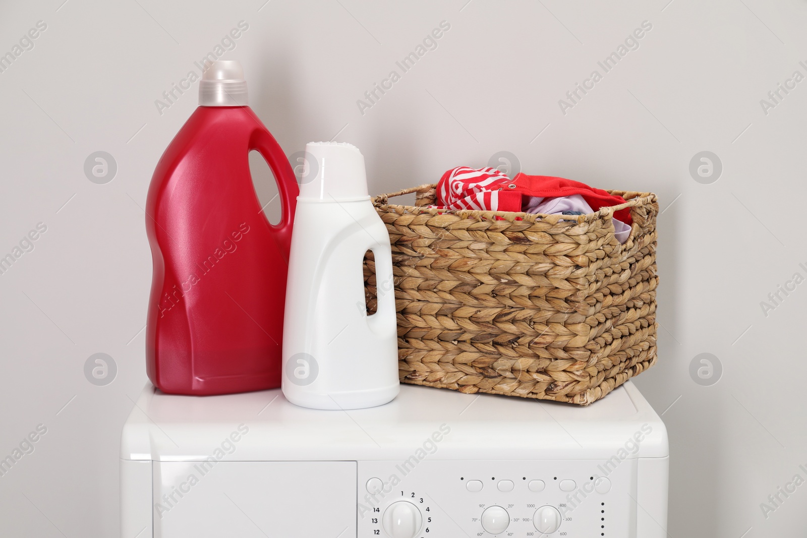 Photo of Baby clothes in wicker basket and laundry detergents on washing machine near light wall