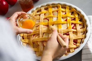 Photo of Woman applying liquid egg onto traditional English apple pie with brush at table, closeup