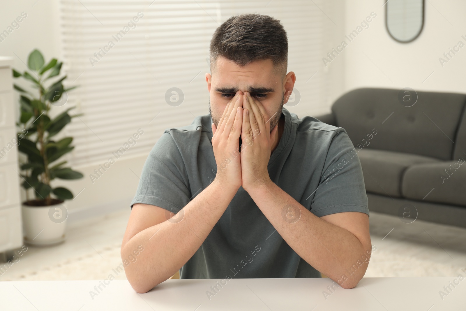 Photo of Sad man covering face with hands at white table indoors