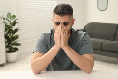 Sad man covering face with hands at white table indoors