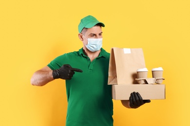 Photo of Courier in medical mask holding packages with takeaway food and drinks on yellow background. Delivery service during quarantine due to Covid-19 outbreak