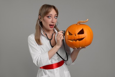 Emotional woman in scary nurse costume with carved pumpkin and stethoscope on light grey background. Halloween celebration