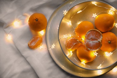 Photo of Fresh ripe tangerines and glowing fairy lights on white cloth, flat lay. Space for text