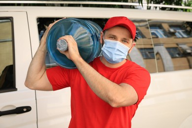 Photo of Courier in medical mask holding bottle of cooler water near car outdoors. Delivery during coronavirus quarantine