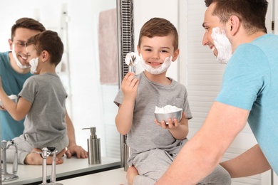 Photo of Little son with shaving brush and bowl full of foam near dad in bathroom