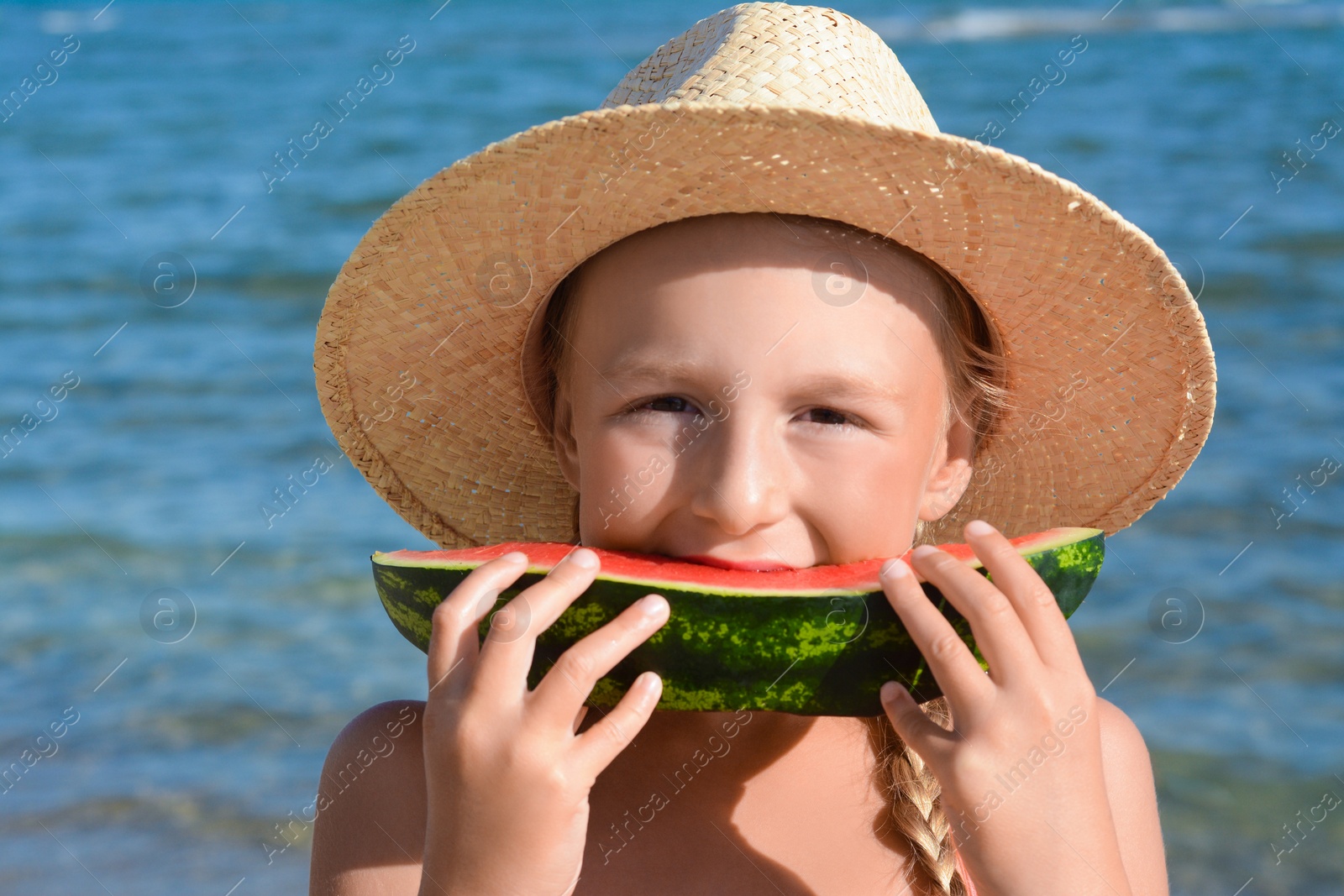 Photo of Cute little girl in straw hat eating juicy watermelon on beach, closeup