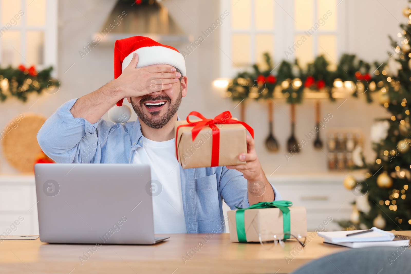 Photo of Celebrating Christmas online with exchanged by mail presents. Man in Santa hat covering eyes before opening gift box during video call on laptop at home