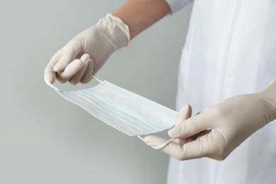 Doctor in medical gloves holding protective mask on light grey background, closeup