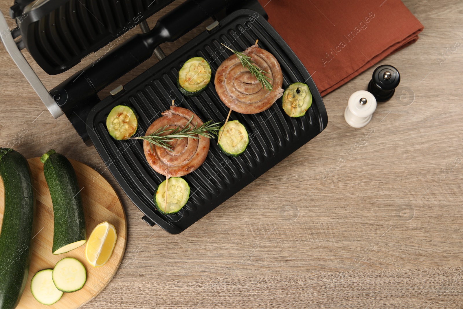 Photo of Electric grill with homemade sausages, zucchini and rosemary on wooden table, flat lay. Space for text