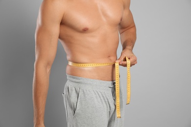 Young man with slim body using measuring tape on grey background, closeup view