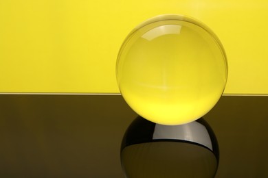 Photo of Transparent glass ball on mirror surface against yellow background, closeup