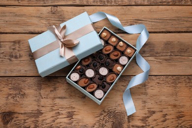 Photo of Open box of delicious chocolate candies and light blue ribbon on wooden table, flat lay