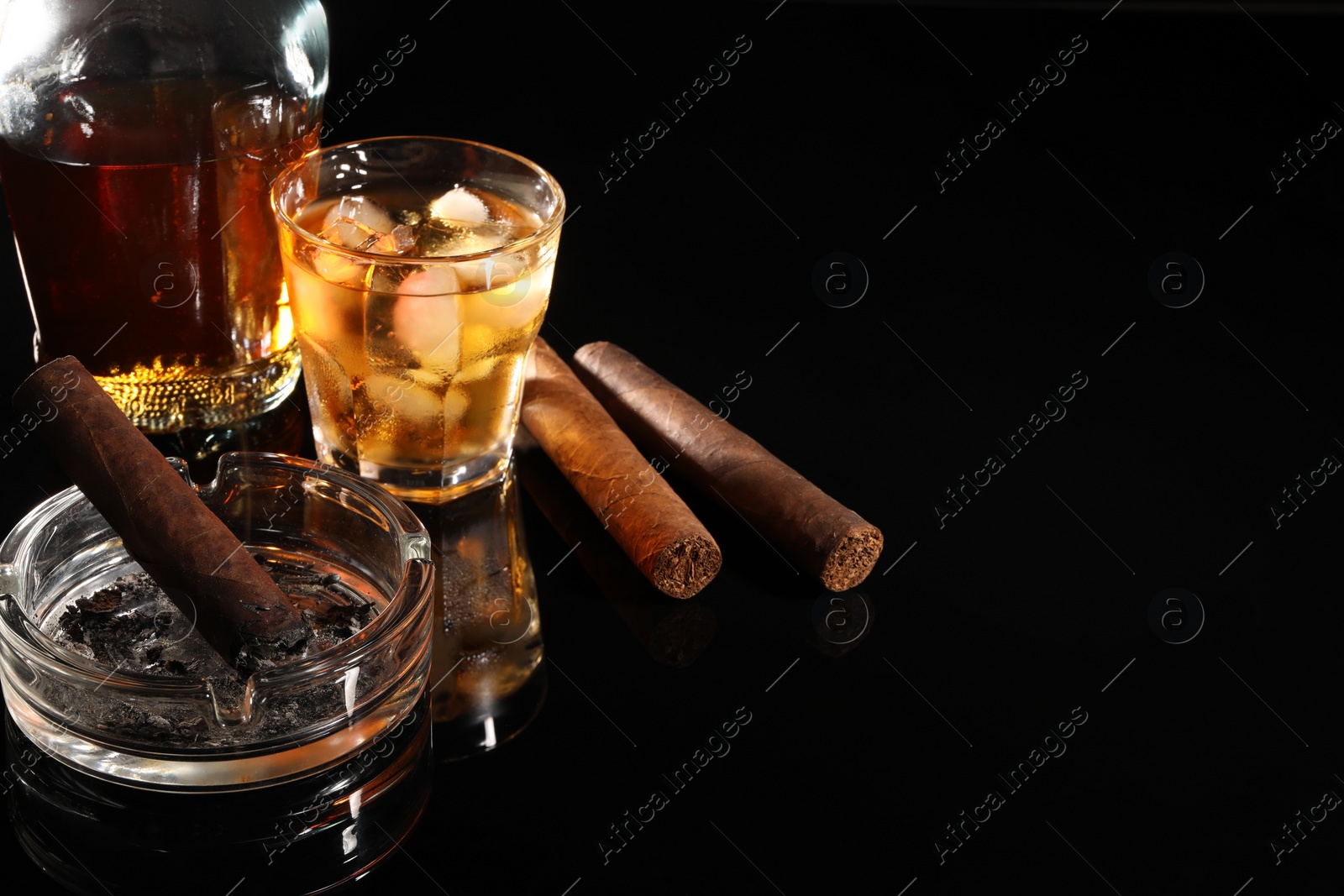 Photo of Smoldering cigar, ashtray and whiskey on black mirror surface. Space for text