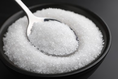 Granulated sugar and spoon in bowl on black table, closeup