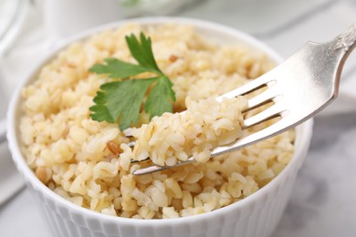 Eating bulgur with fork from bowl on white table, closeup
