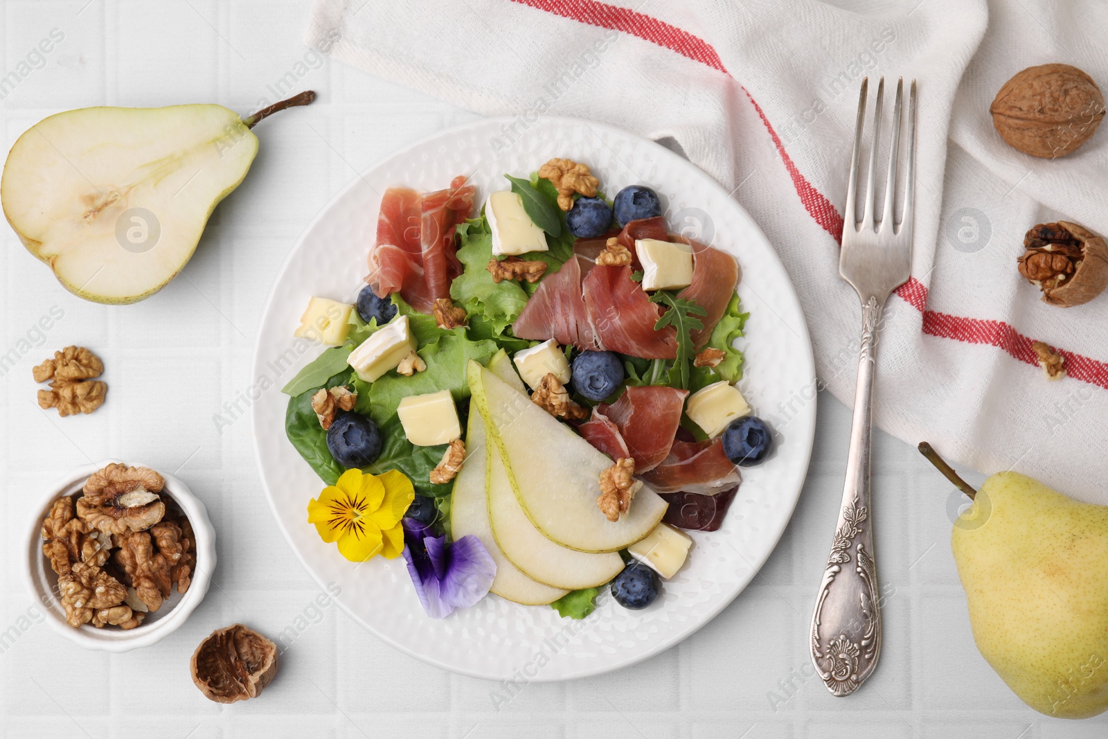 Photo of Tasty salad with brie cheese, prosciutto, walnuts and pear served on white tiled table, flat lay